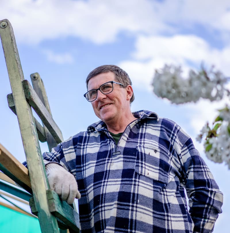 Smiling tradesman in a plaid shirt and work gloves standing by a wooden ladder on a sunny day